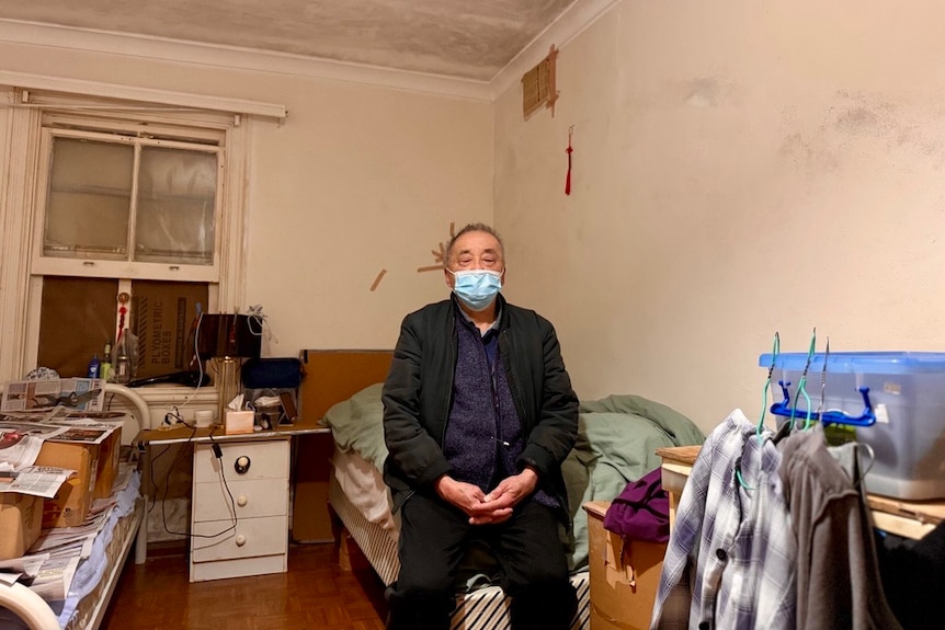 A man in a surgical mask sitting on the edge of a bed in a bedroom.