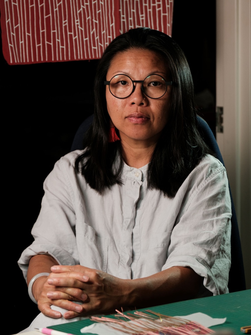 A woman with long black hair and black-framed glasses, wearing a white shirt, sitting with her hands clasped.