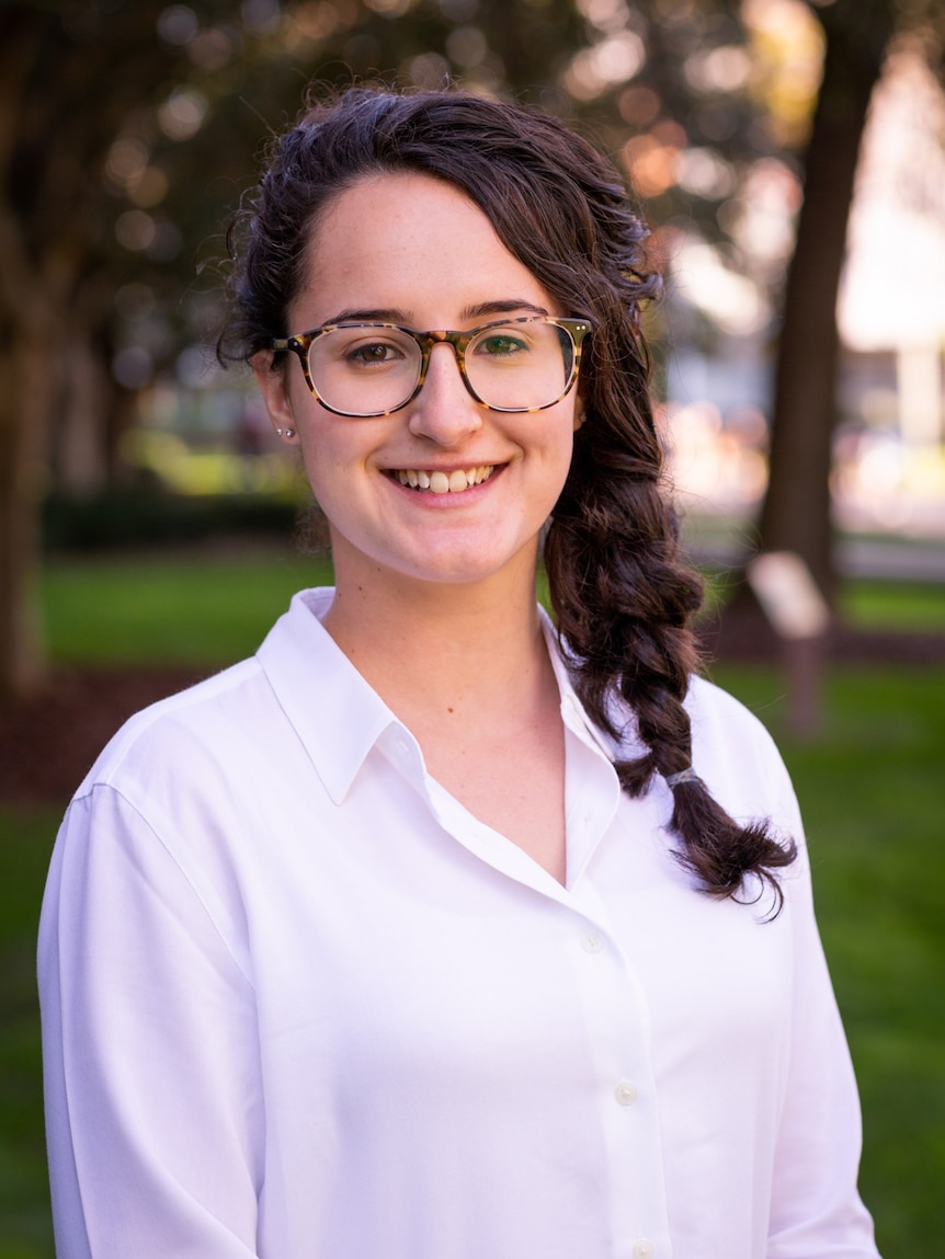 A woman smiles. She wears glasses and a white shirt and her long brown hair is in a side plait.
