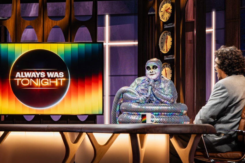 A TV still of Megan Wilding and Tony Armstrong, both mid-30s, Indigenous, in a TV studio, Wilding dressed as a silver snake.