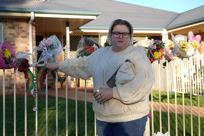 A middle-aged woman in glasses and a jumper touches a tribute left on a fence in front of a suburban home.
