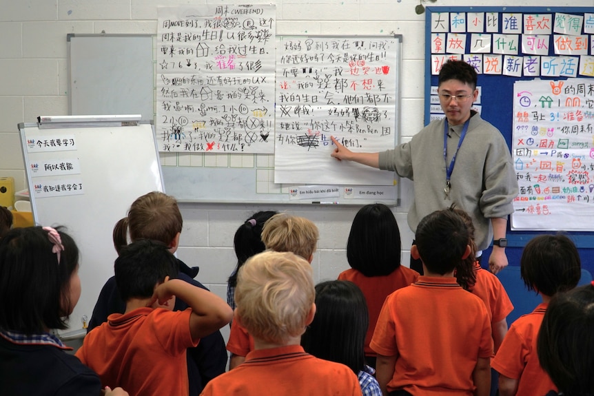 A teacher stands at the front of the classroom in front of big boards covered in Chinese characters.