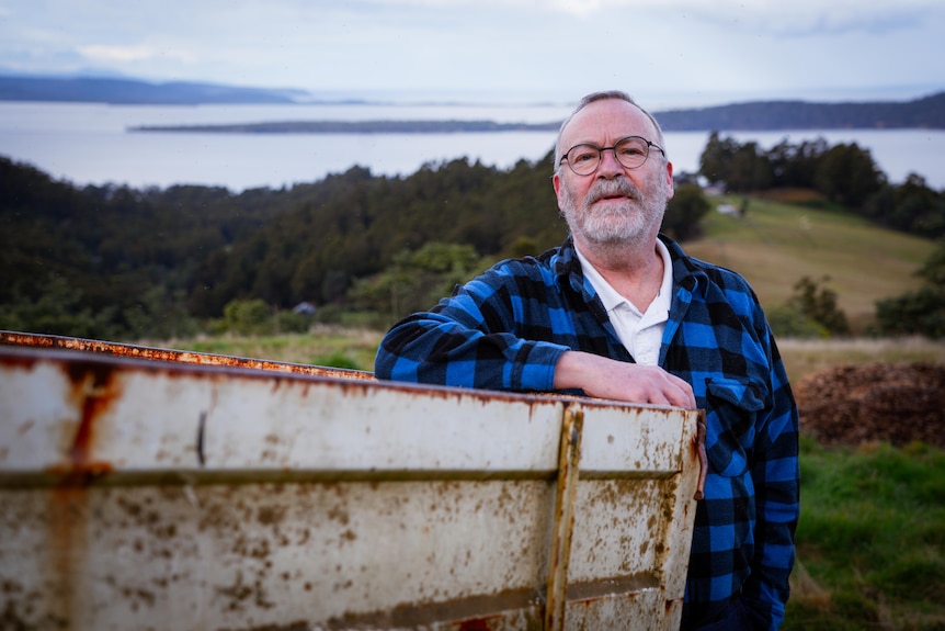 A middle-aged, bearded man in a checked shirt stands on a farm overlooking the sea.