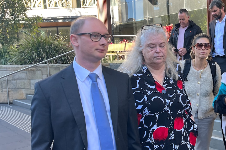 A man in a dark suit and an older woman with long, grey hair walk away from a court building.