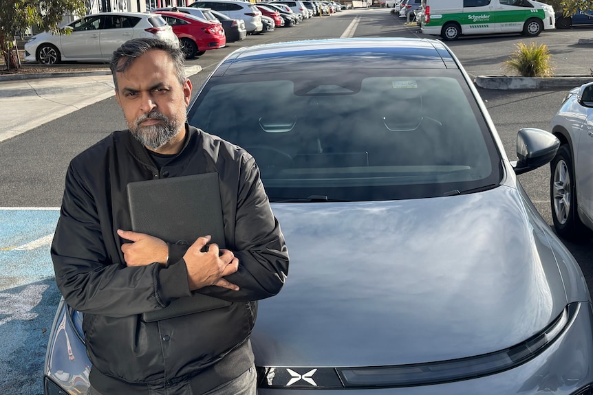 A middle-aged, bearded man clutches a laptop while leaning on a dark car.