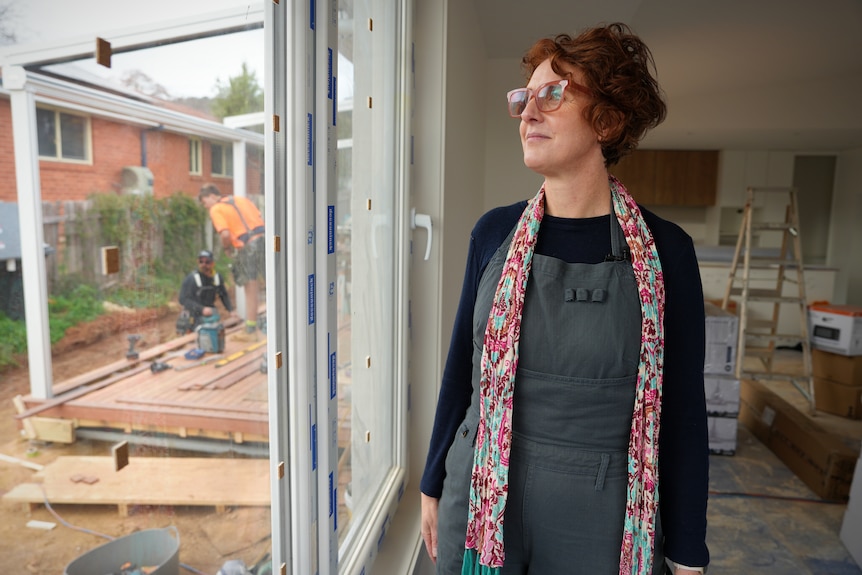 A woman wearing glasses and a scarf stands inside a home under construction.