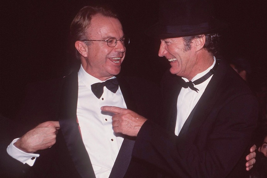 Two men dressed in black suits smile big, one pointing at the other
