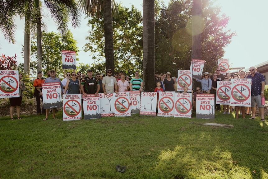 A group of people stand in the grass holding anti-wind farm signs.