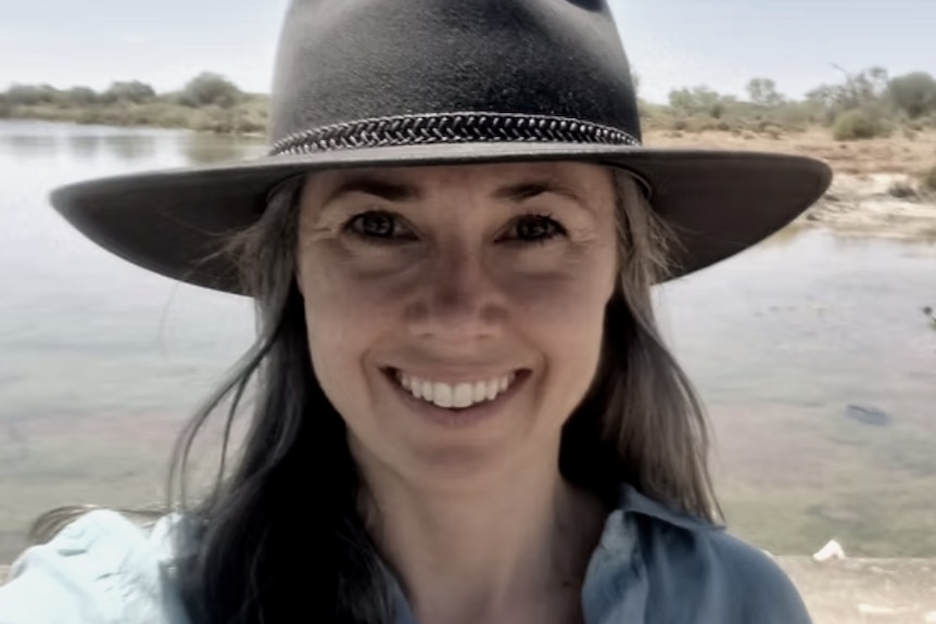 A woman in a wide-brimmed hat smiles in a selfie taken in the sun, in front of an inland pool of water in arid bushland.