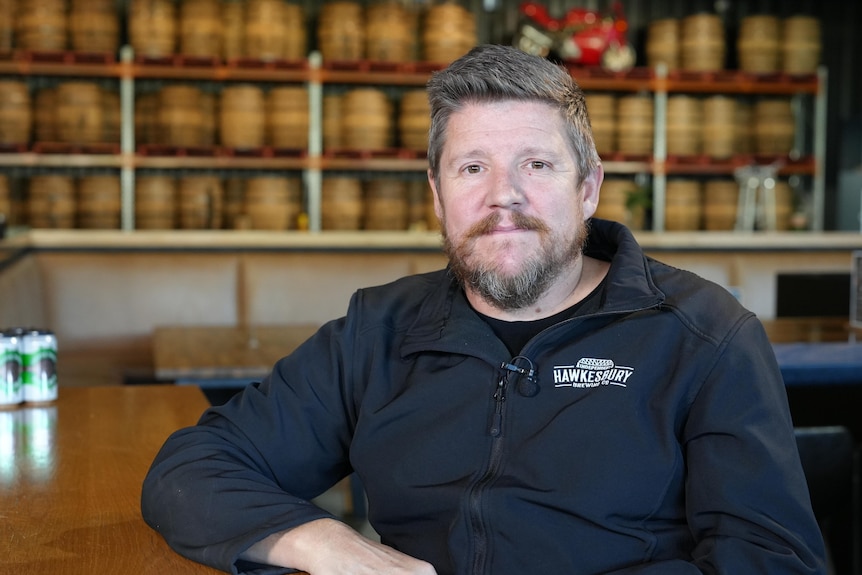 A man with brown hair, wearing a black jacket and sitting at a tall table. Large wooden barrels cover the wall behind him.