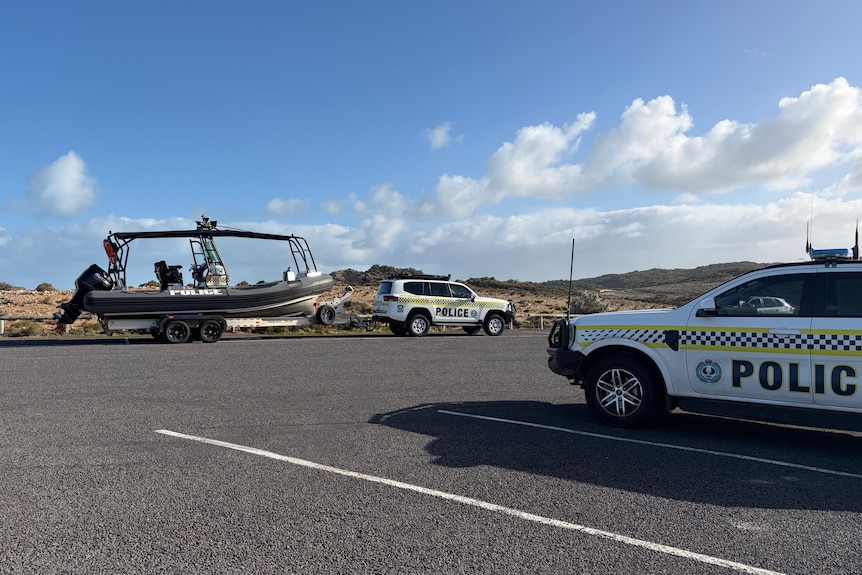A police car towing a search boat in a car park near a rocky cliff