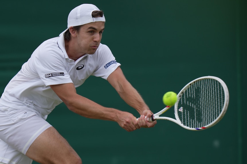 A man in a white cap and white tennis clothes clutches his breath as he takes a shot at a yellow tennis ball