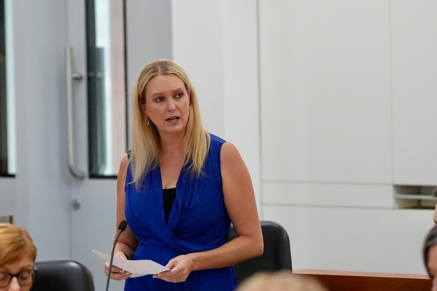 A woman with blonde hair stands in a room with red seats looking serious.