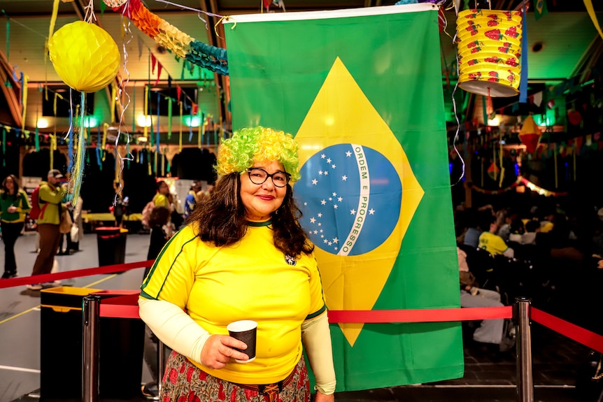 Woman wearing yellow Brazil shirt with yellow and green wig stands in front of a Brazillian flag with a lively event behind her
