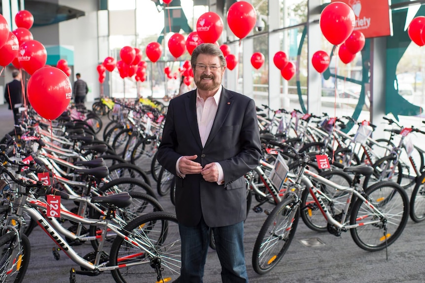 A dark haired man with a beard and glasses in a dark suit stands rows of bicycles with red balloons attached to them.