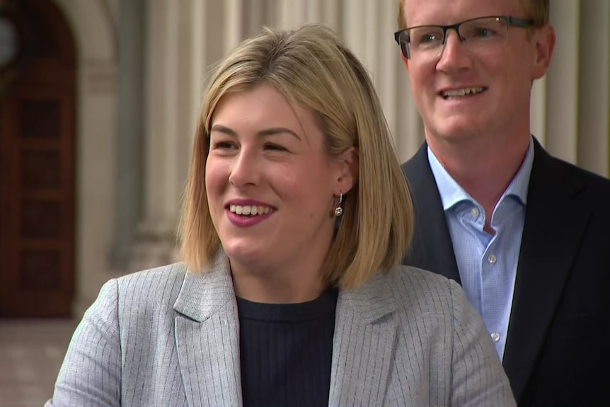 A blonde haired woman and a red headed man with glasses, both in blazers, look off camera, smiling.