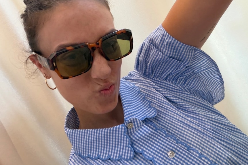 Mikaela poses with sunglasses and a blue check collared button up shirt.