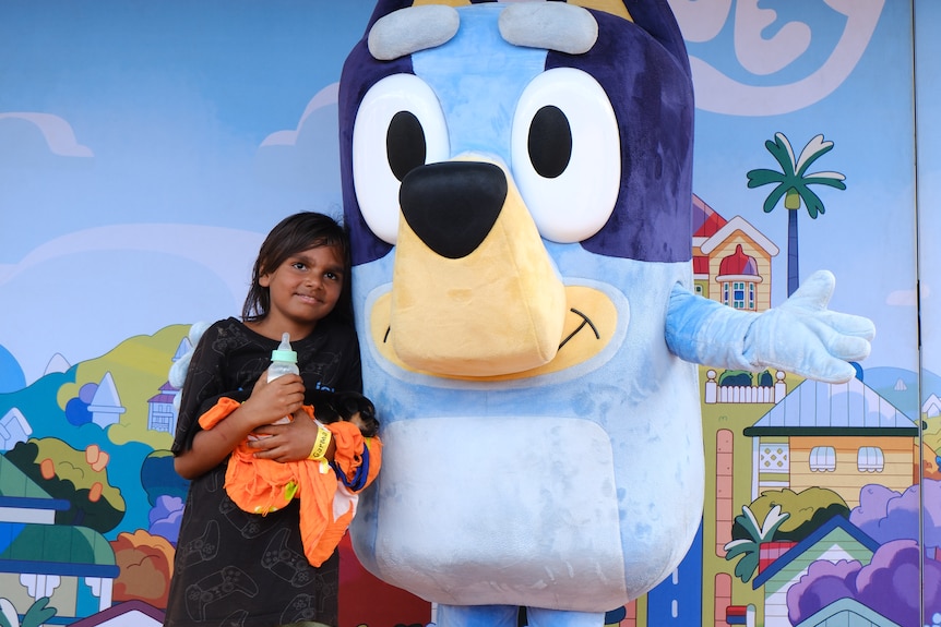 A smiling, dark-haired boy holds a puppy as he cuddles up to a person in a Bluey suit.