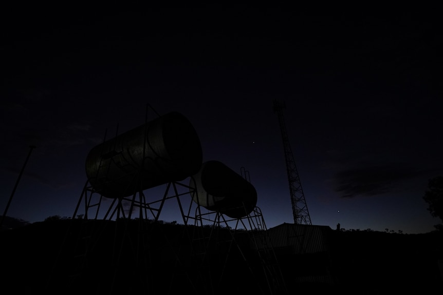 Metal structures at a remote roadhouse and rocky landscapes are silhouetted against the darkening night sky behind.