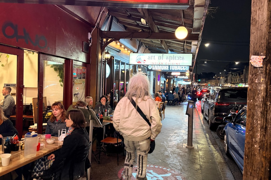 Man with white hair walking as people eat and drink on street at night