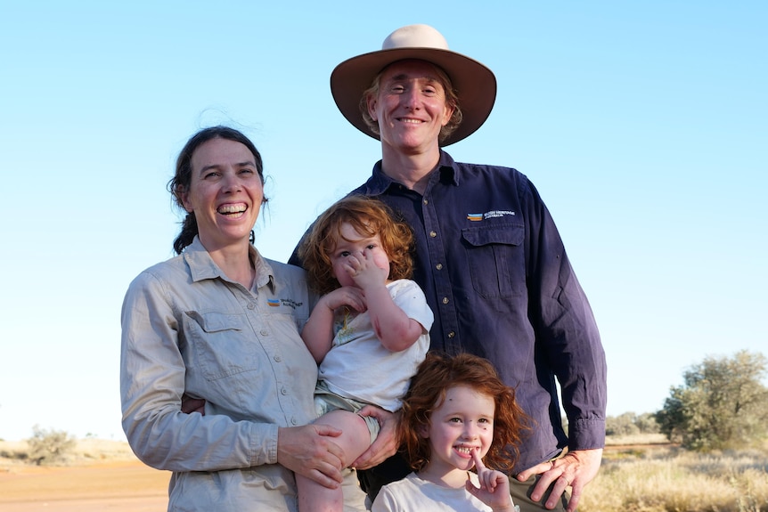 A woman holds a little girl standing beside a man and another child. Flies sorround the family.