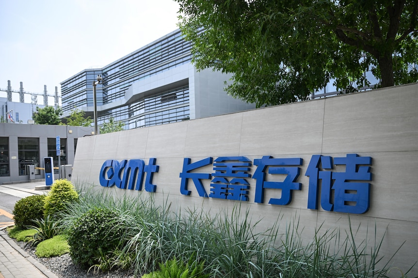 A sign with a blue logo saying "CXMT" and with some Chinese characters, out front of a large building.