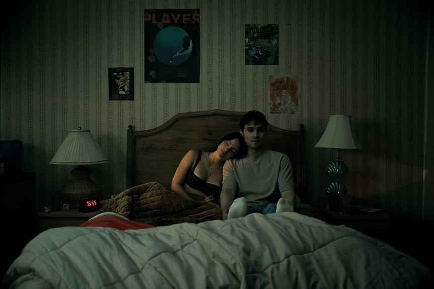 A young man and woman sit on a bed in a dark room, the woman has her head on the man's shoulder