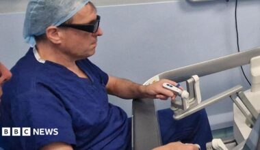 A medic wearing blue surgical scrubs and a head covering is sitting at a screen which shows an operation. He is holding two controls to direct the operation.