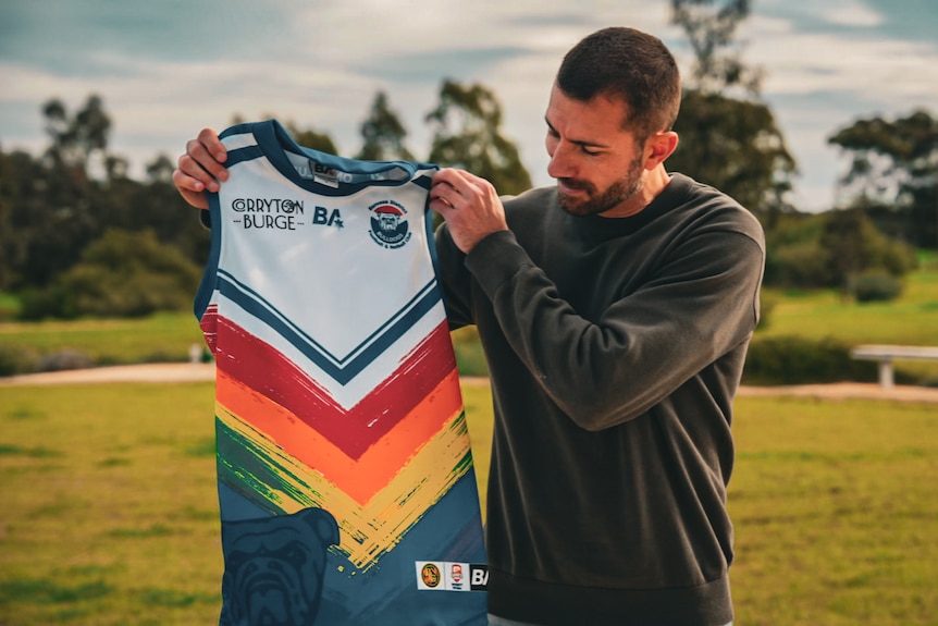 A man holds up an Australian Rules Football guernsey, featuring a white base with rainbow-coloured chevrons