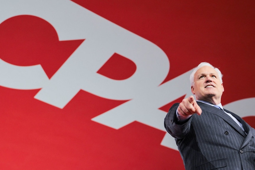 A man in a suit looking on and pointing. A large sign saying CPAC is visible behind him.