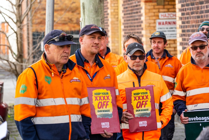 workers in orange jumpers outside an old red brick building holding signs petitioning for their brewery to not close down