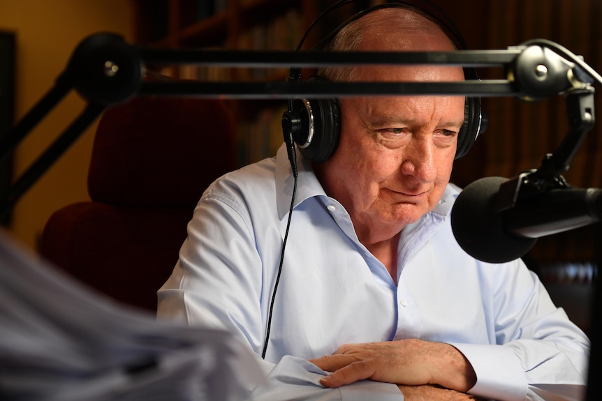 Alan Jones sitting at his desk, in a blue work shirt, with headphones on and a microphone near his mouth.