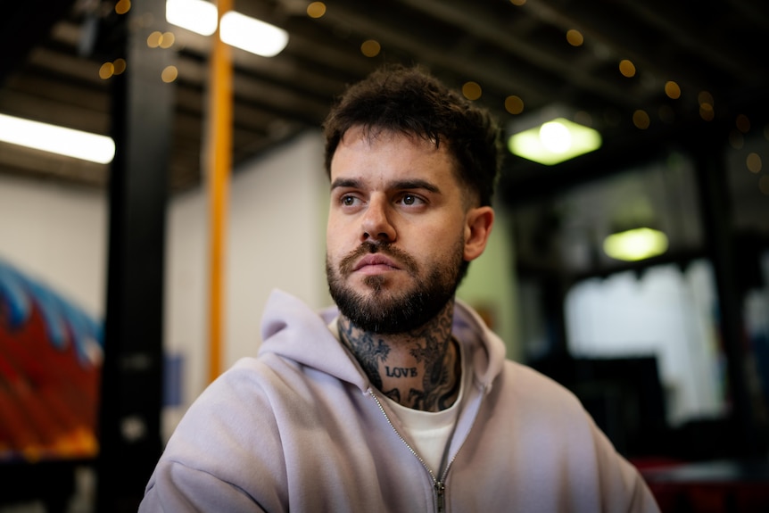 Close up of Kieran wearing a purple zip up hoodie that covers part of his neck tattoos, green lights reflect on the wall.