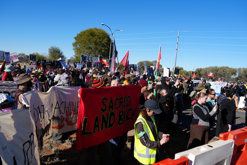 A large group of  protesters gathered on a wide road, on one side of a roadblock.