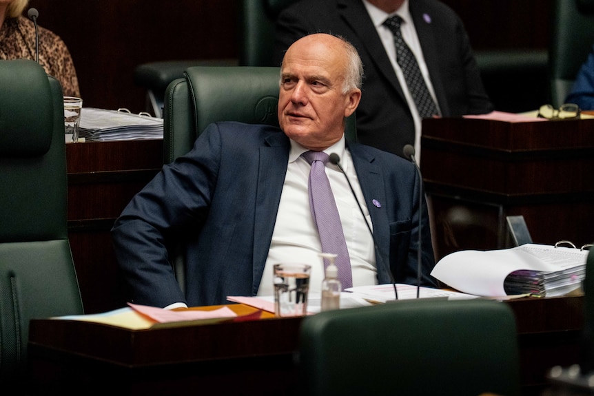 A man dressed formally sits in a green chair in parliament among others