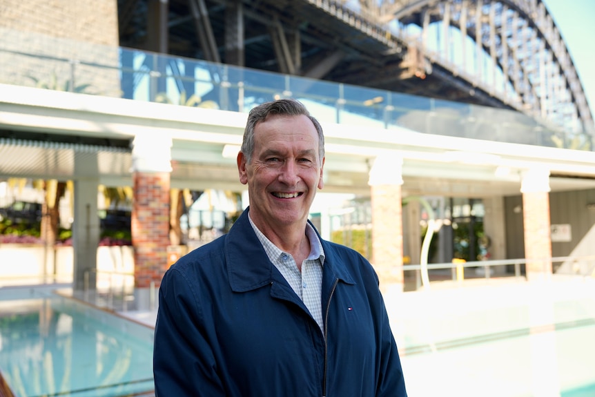 Graham Windeatt smiles for a photo at a swimming pool with the Sydney Harbour Bridge in the background.