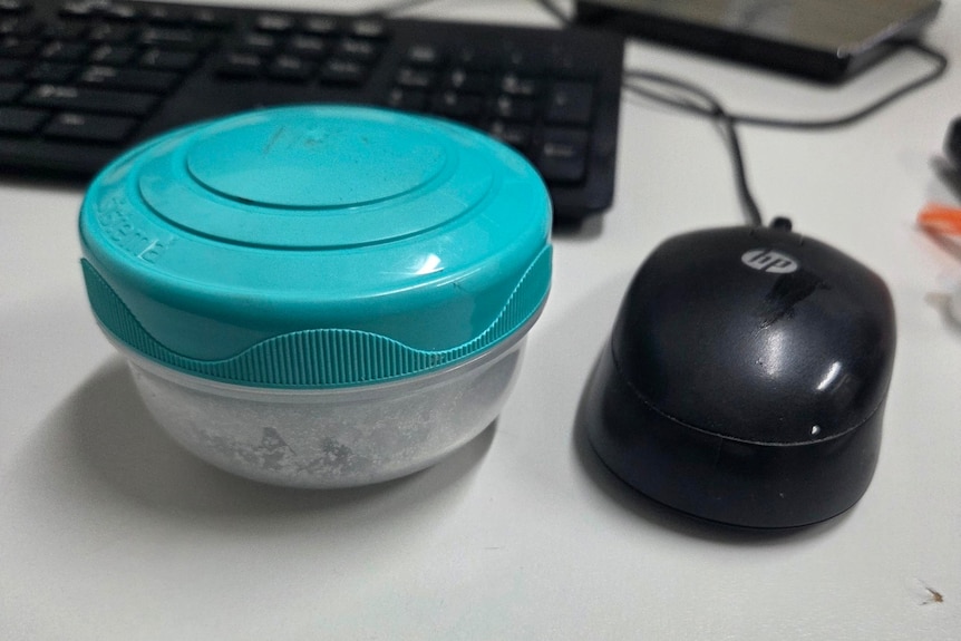 A clear container with a blue lid containing a white powder on a desk next to a mouse and keyboard.