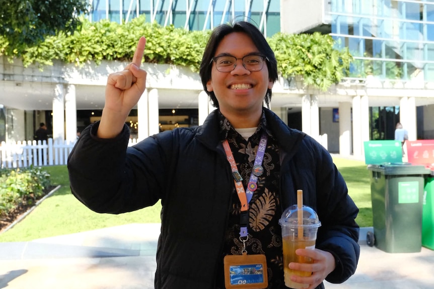A young Indonesian man, wearing glasses, smiles widely and holds up a finger and a drink.
