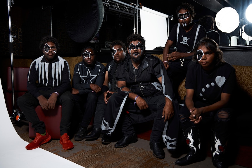 Six members of Mulga Bore Hard Rock in face paint and matching black outfits pose on a couch in a photo studio