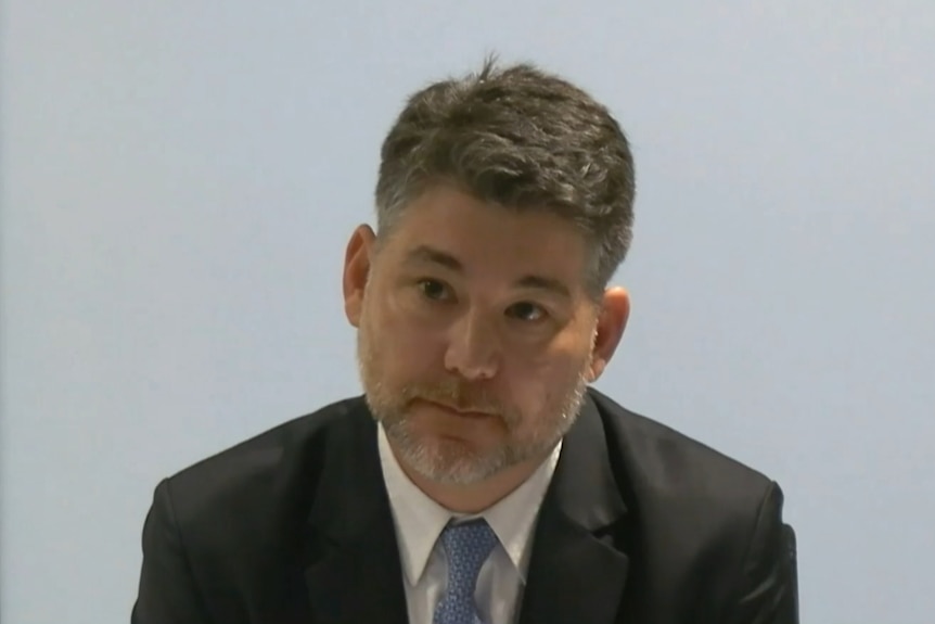 Mr Fang wearing a black suit and blue tie sitting in a chair looking serious.
