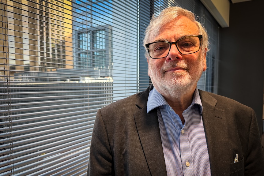 A white haired man with a beard, dark rimmed glasses, grey jacket and purple and white striped shirt stands near white blinds.