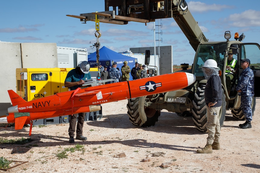 A red missile is lowered on a crane as people inspect it.