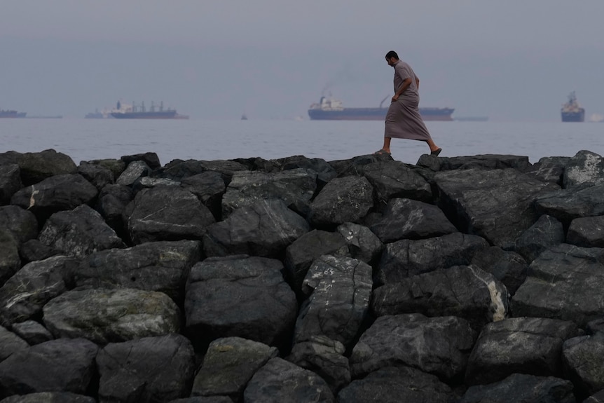 A man walking along a rock seawall with large oil tankers lined up in the background.