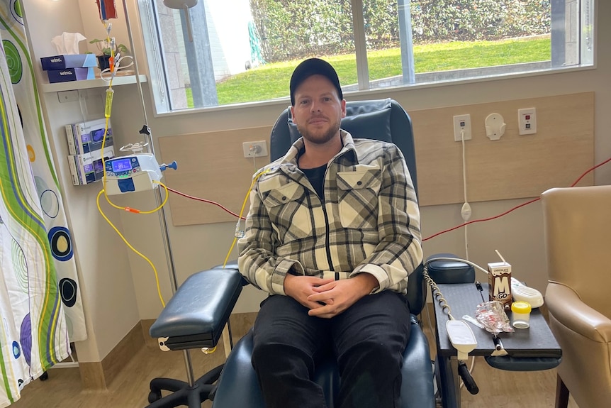 A young man wearing a cap sits in a chair getting chemo.