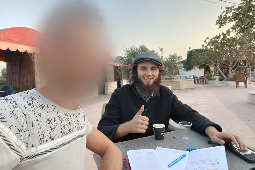 A young man with a beard gives a thumbs up gesture while posing with another man, who is blurred.