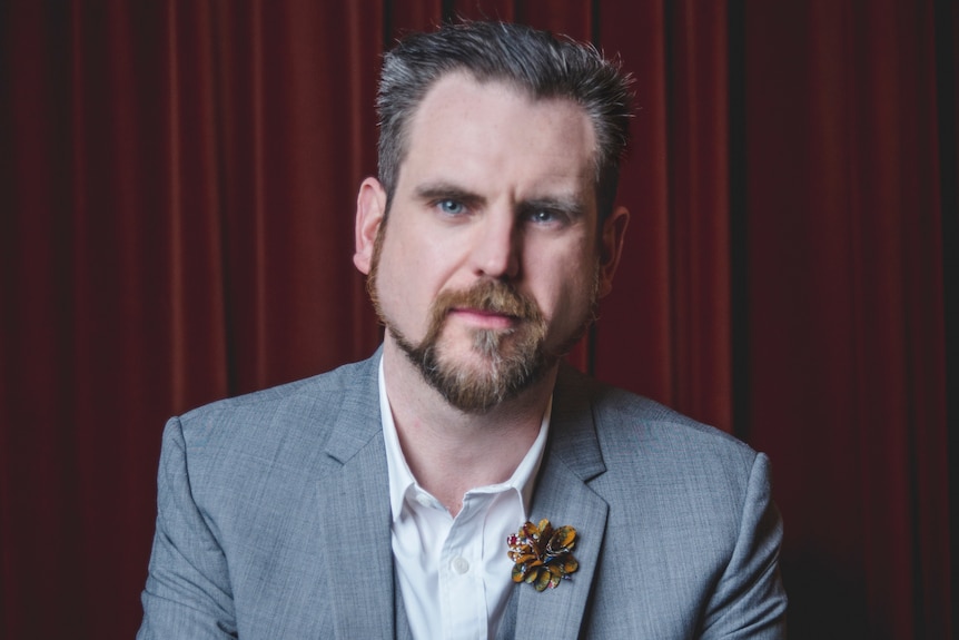 Zak Hepburn with beard and moustache, brown hair, grey suit jacket, white shirt