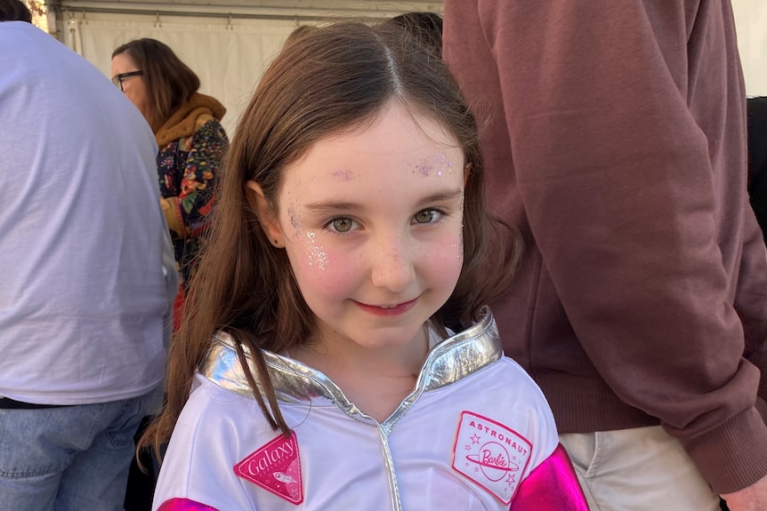 A young girl in a pink and white Barbie astronaut suit looks at the camera smiling