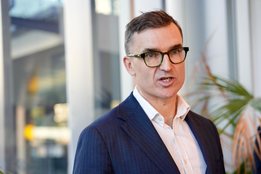 A tight head and shoulders shot of WA Housing Minister John Carey speaking at a media conference, in a suit and spectacles.