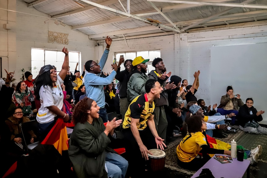 A room full of people, some wearing red, green and yellow, clap and raise their arms as they look forward.