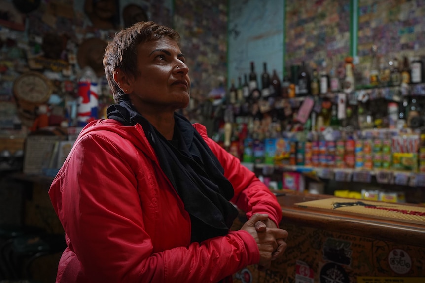 A woman sitting at the bar inside a dim hotel, looking over the mementos plastered on the walls around her.